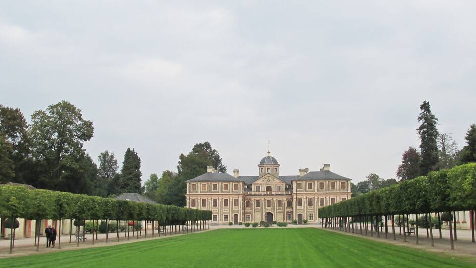 A large, classical building with a symmetrical facade, surrounded by a well-manicured lawn and rows of trees.