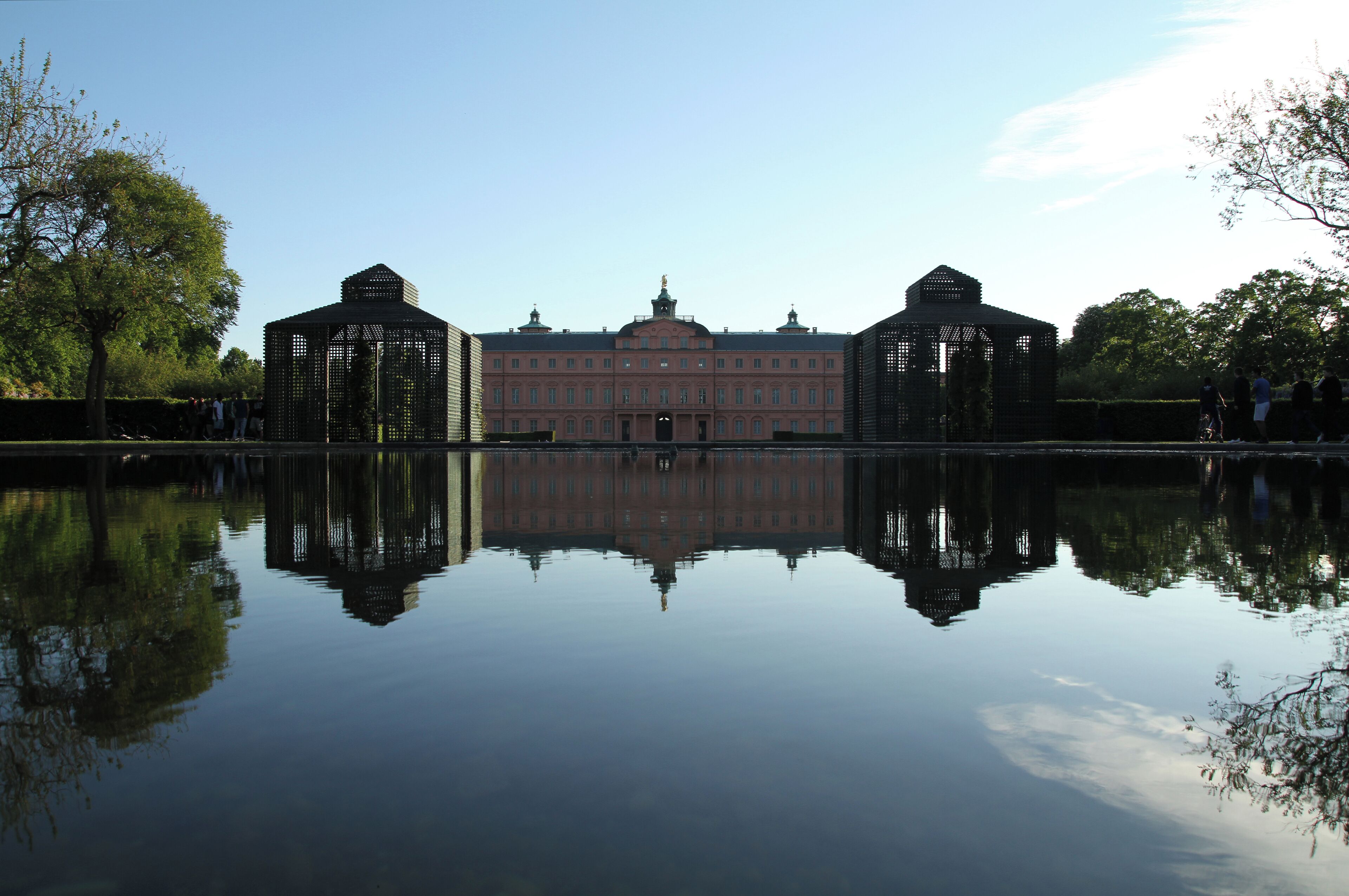 A large building with a reflection in the water, surrounded by trees and a clear sky.