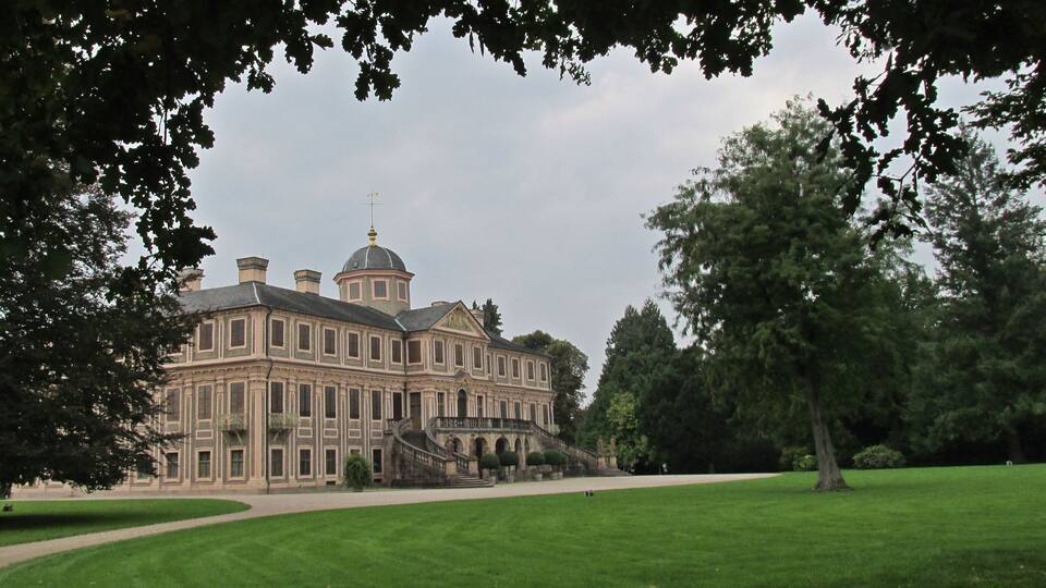 A large, historic building with a dome and symmetrical architecture, surrounded by a well-maintained lawn and trees.