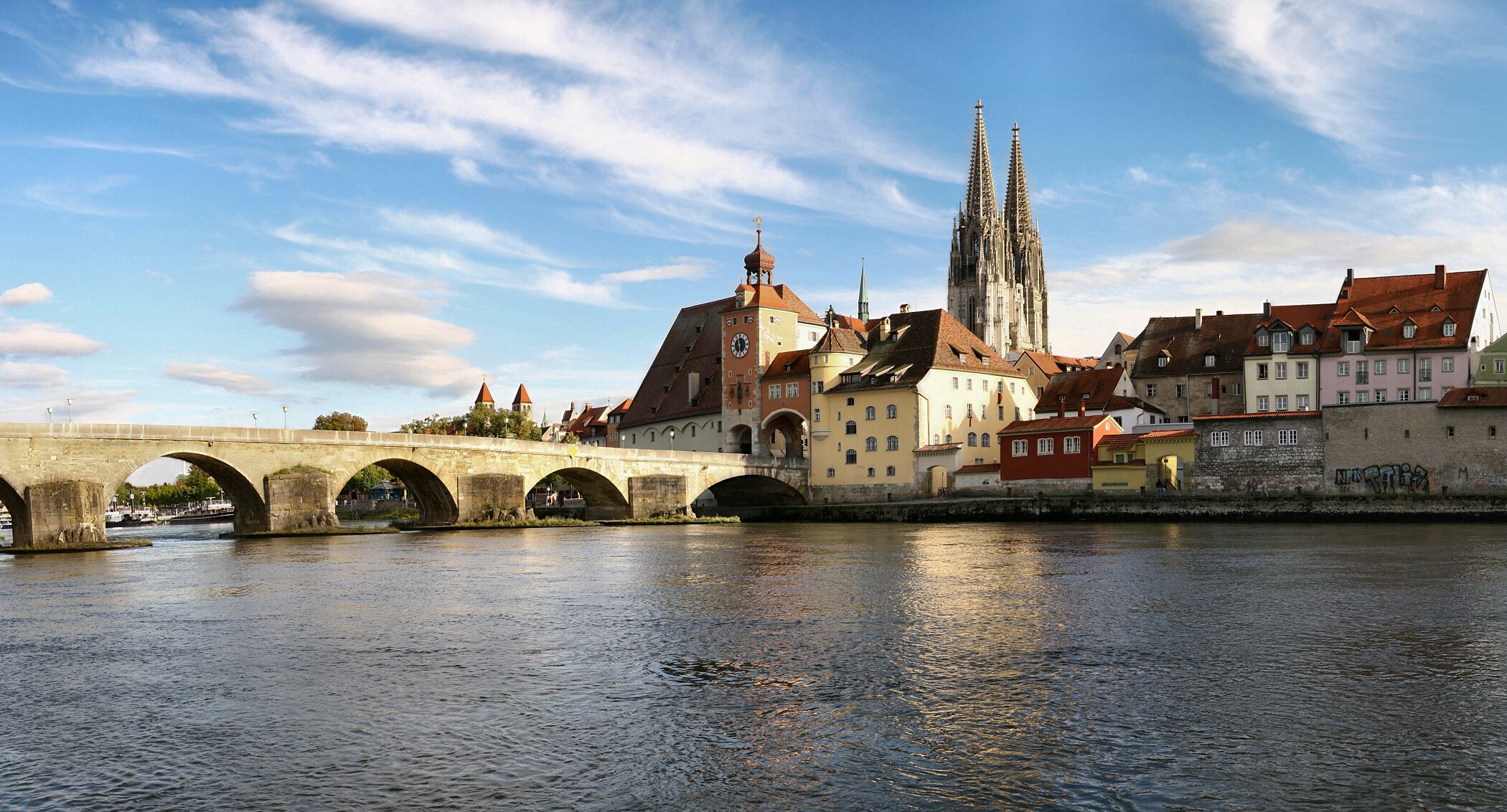 Eine historische Brücke mit Bögen, ein Dom mit zwei Türmen und bunte Häuser entlang eines Flusses.