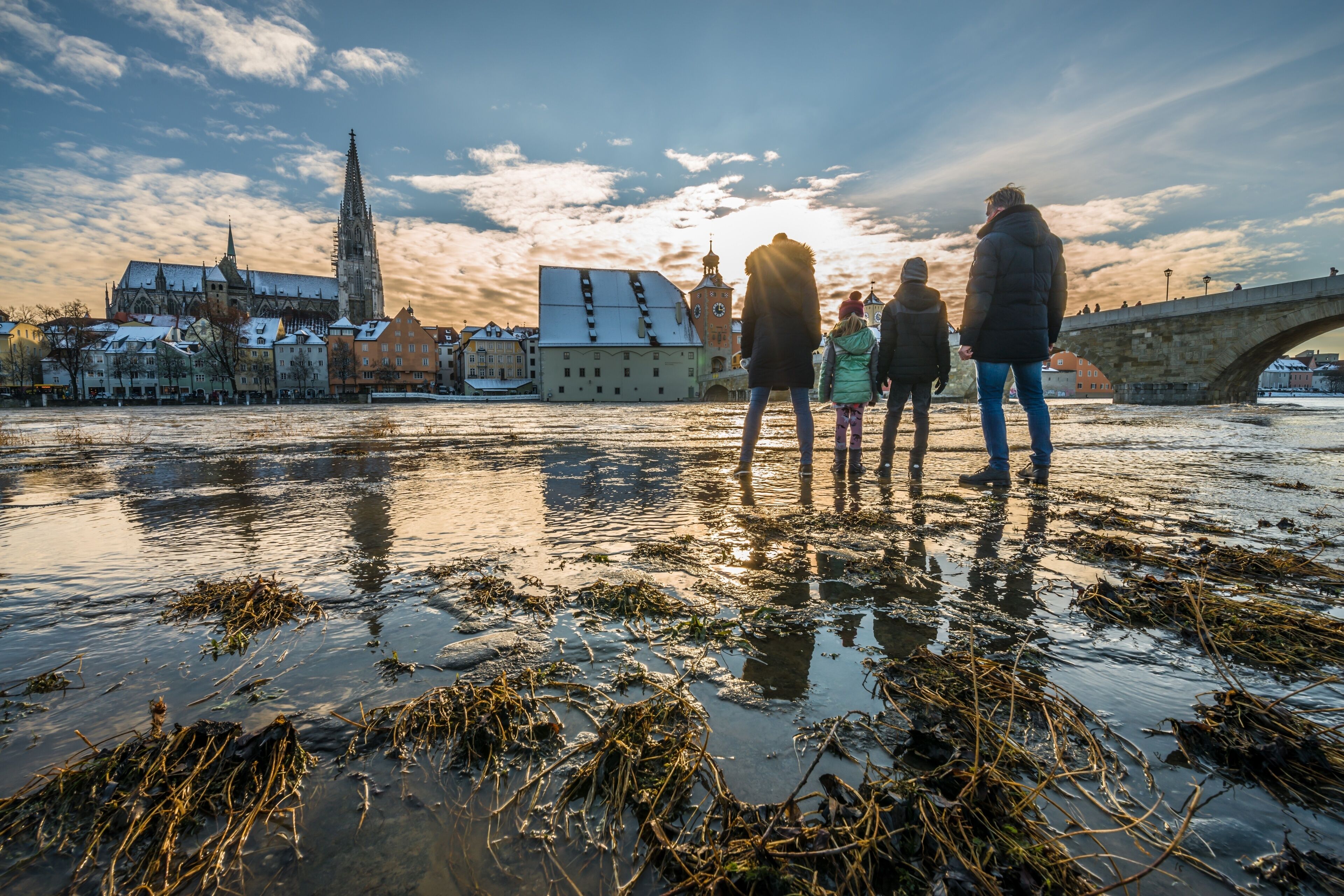 A group of people standing on a frozen riverbank with a historic church and a stone bridge in the background.