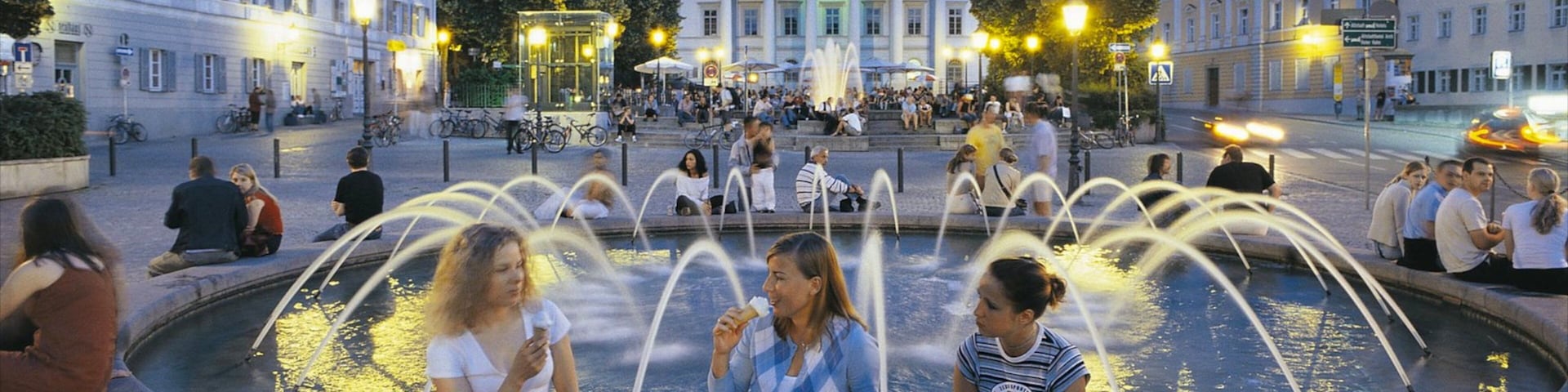 A public square with a fountain, people sitting around it, and classical architecture in the background.