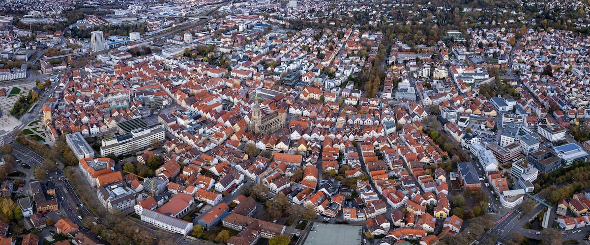 An aerial view of a densely packed urban area with red-roofed buildings, a prominent church tower, and a mix of modern and traditional architecture.