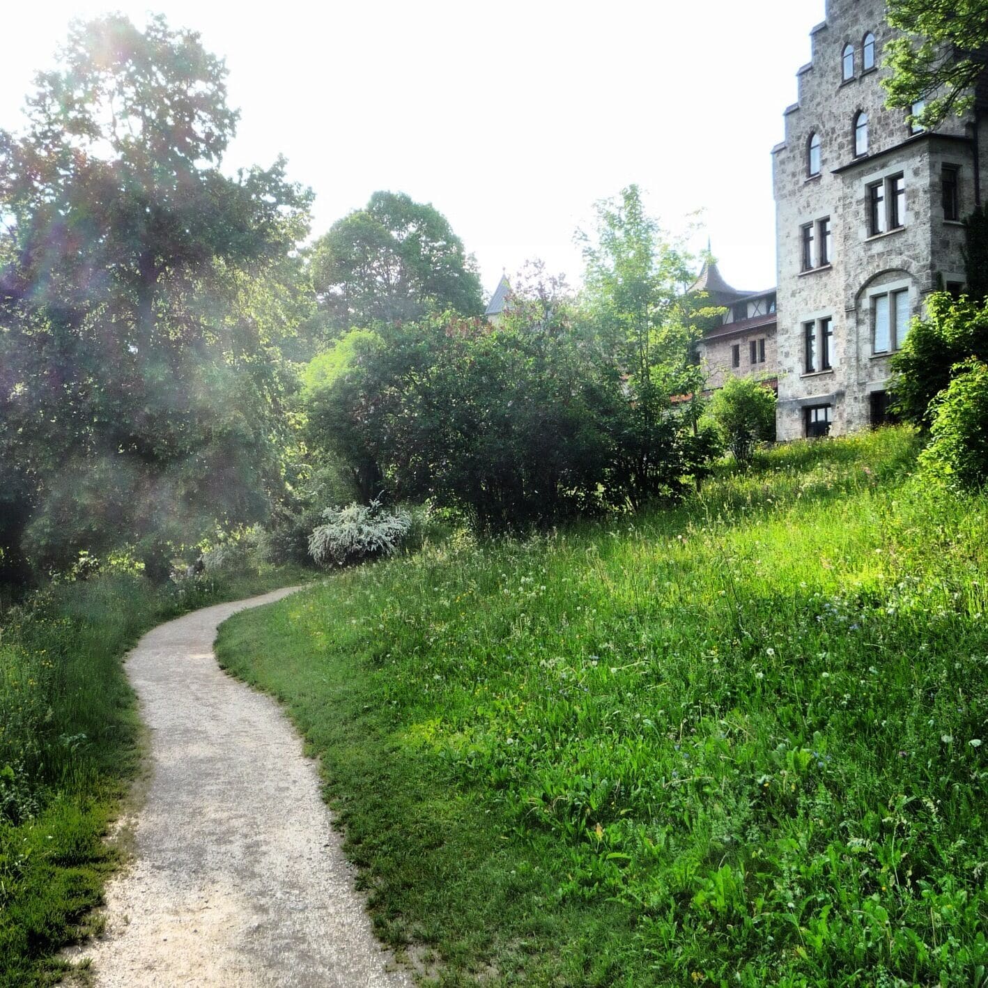 A gravel path winding through a lush green landscape with dense foliage and a historic stone building on the right.