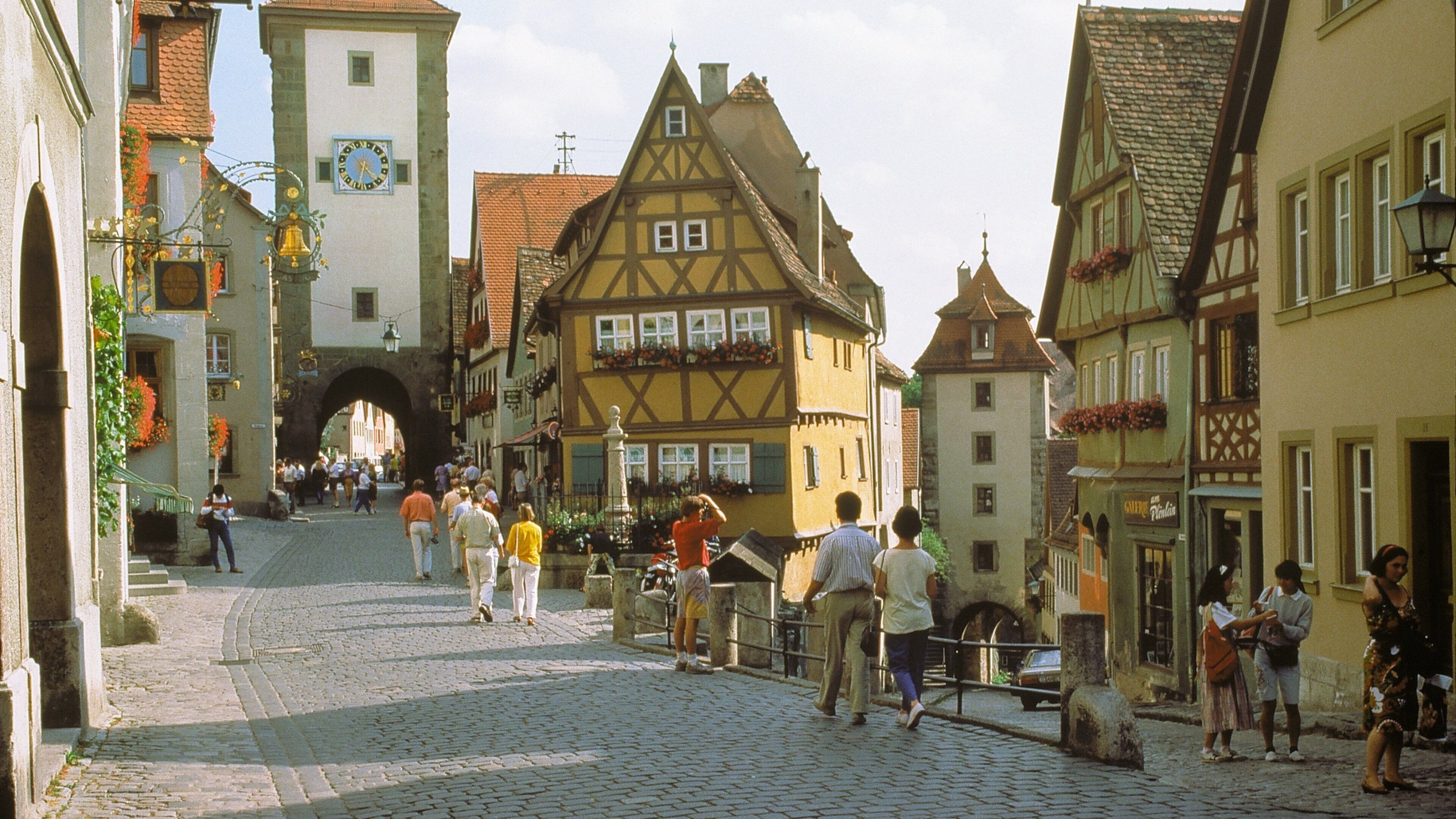 A cobblestone street lined with historic half-timbered buildings, a clock tower, and a stone archway.