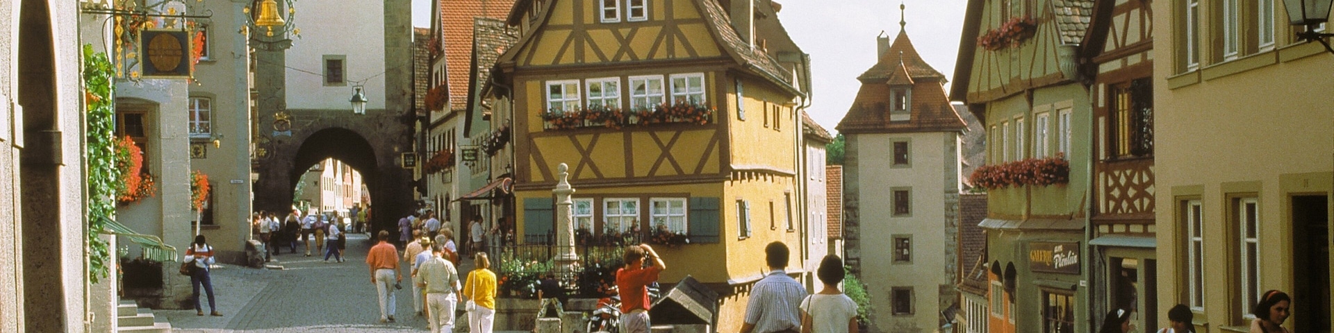 A cobblestone street lined with historic half-timbered buildings, a clock tower, and a stone archway.