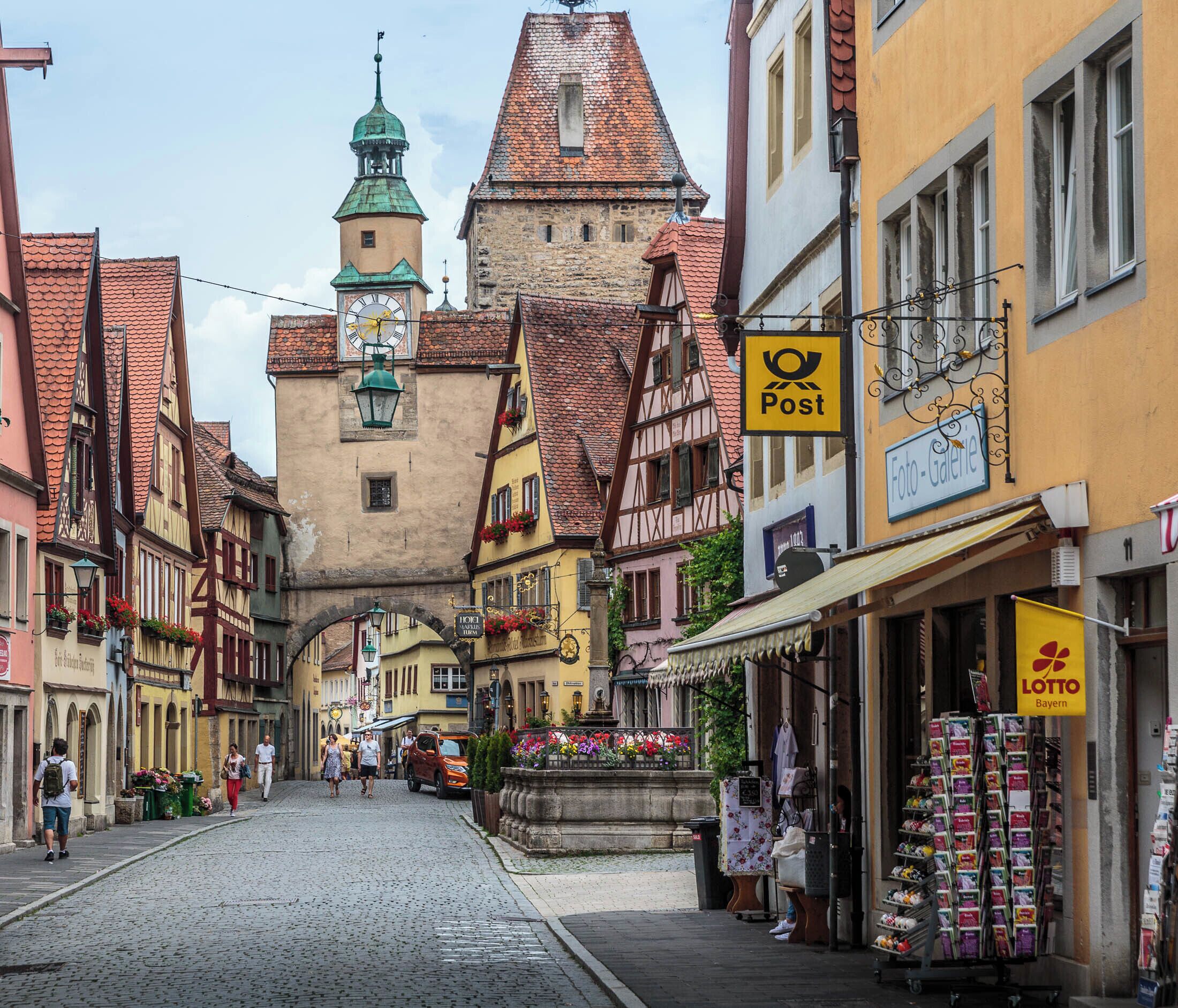 A cobblestone street lined with traditional half-timbered houses, a church tower, and various shops including a lottery and a photo gallery.