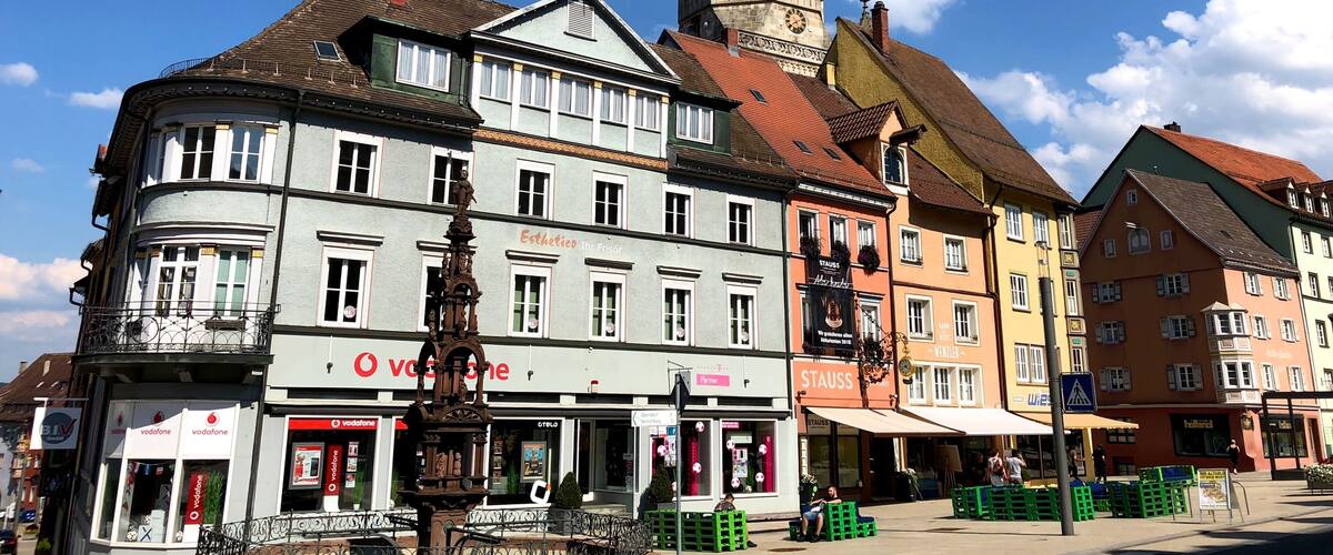 A historic town square with a prominent church tower, colorful buildings, and a street with shops and cafes.