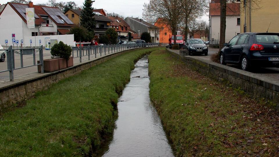 A narrow water channel flanked by grass and buildings, with parked cars and a pedestrian path.
