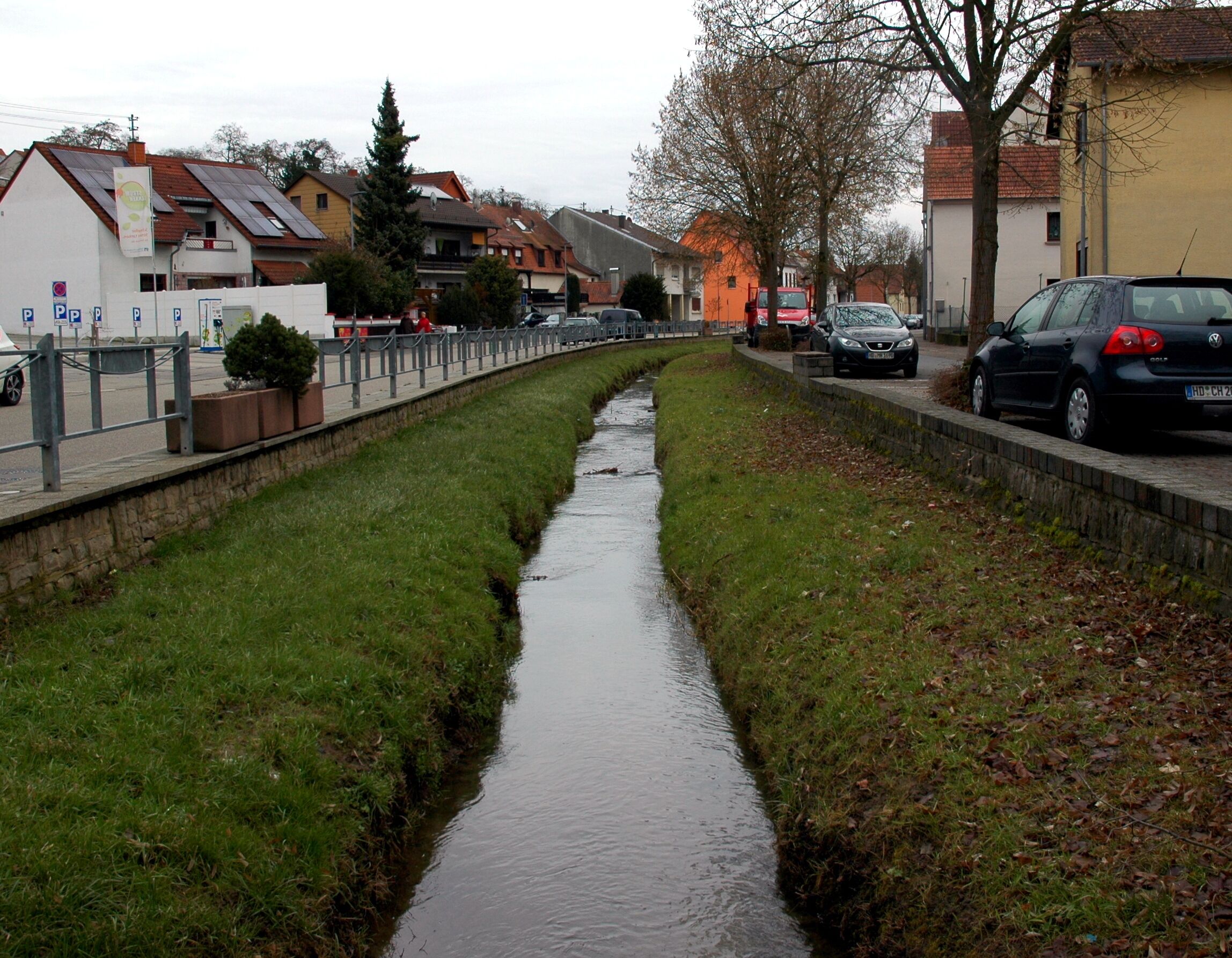 A narrow water channel flanked by grass and buildings, with parked cars and a pedestrian path.