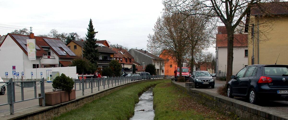 A narrow water channel flanked by grass and buildings, with parked cars and a pedestrian path.
