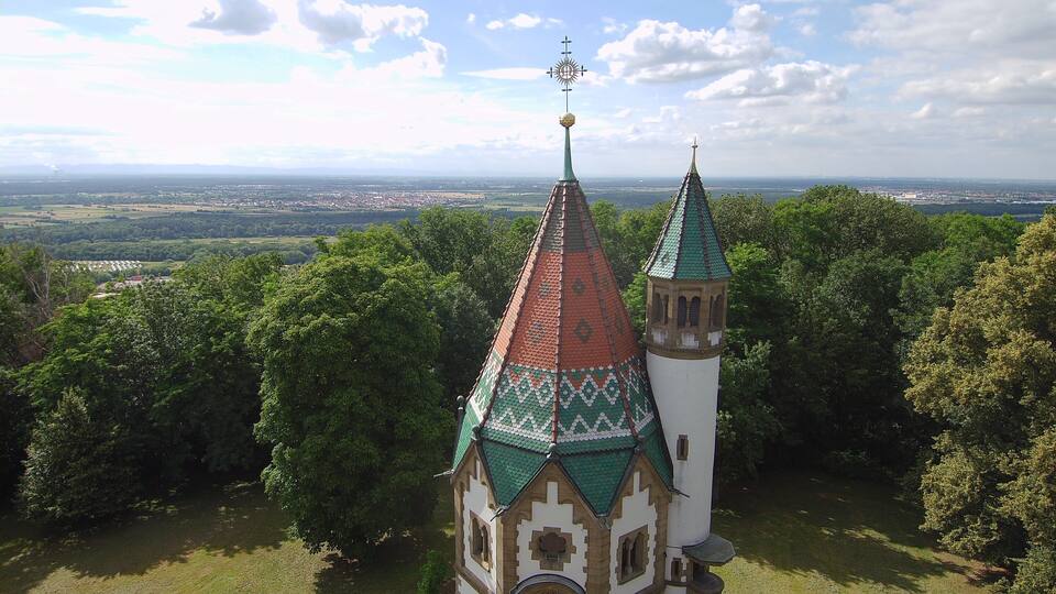 A church with a green roof and a weather vane, surrounded by trees and a distant cityscape.