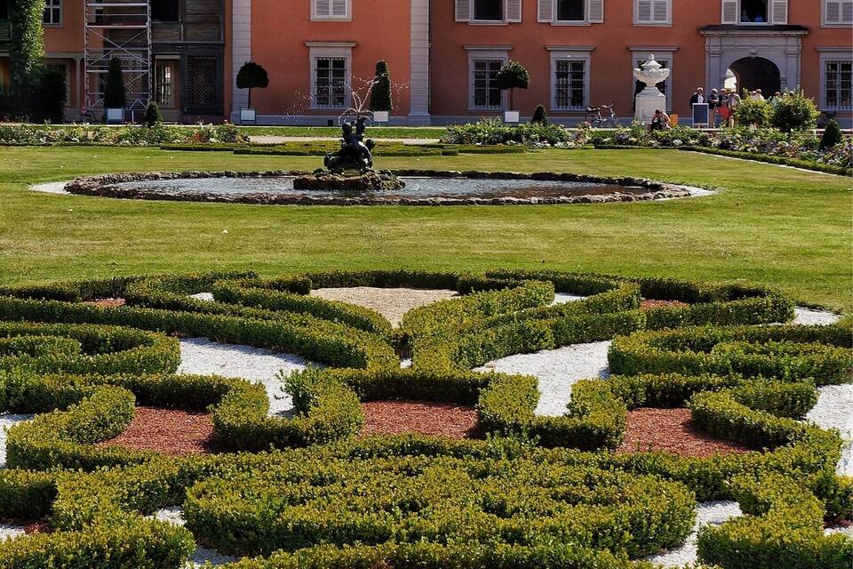 A historic building with a clock tower, surrounded by a formal garden with geometric hedges and a central fountain.