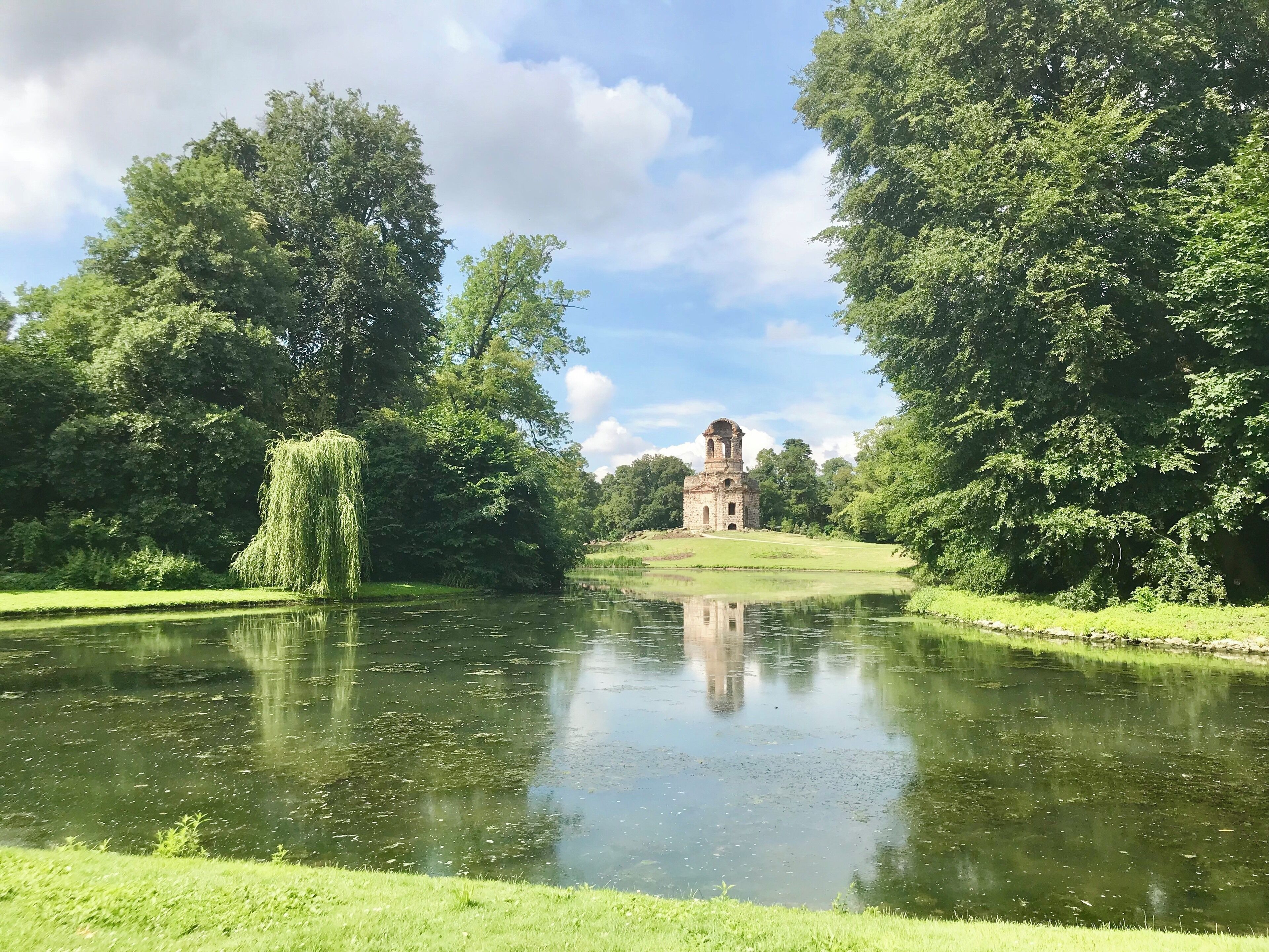 A serene pond with a weeping willow tree, lush greenery, and a historic stone tower in the distance.