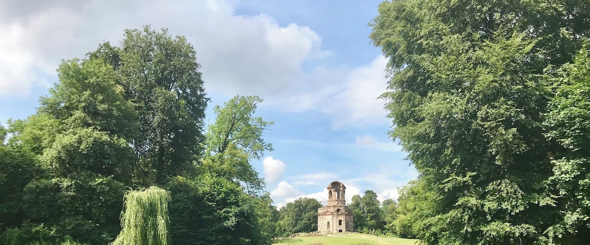 A serene pond with a weeping willow tree, lush greenery, and a historic stone tower in the distance.