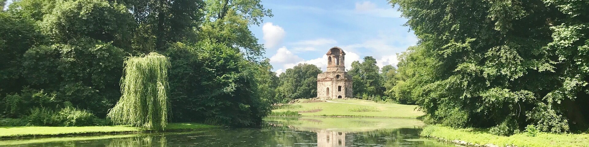 A serene pond with a weeping willow tree, lush greenery, and a historic stone tower in the distance.