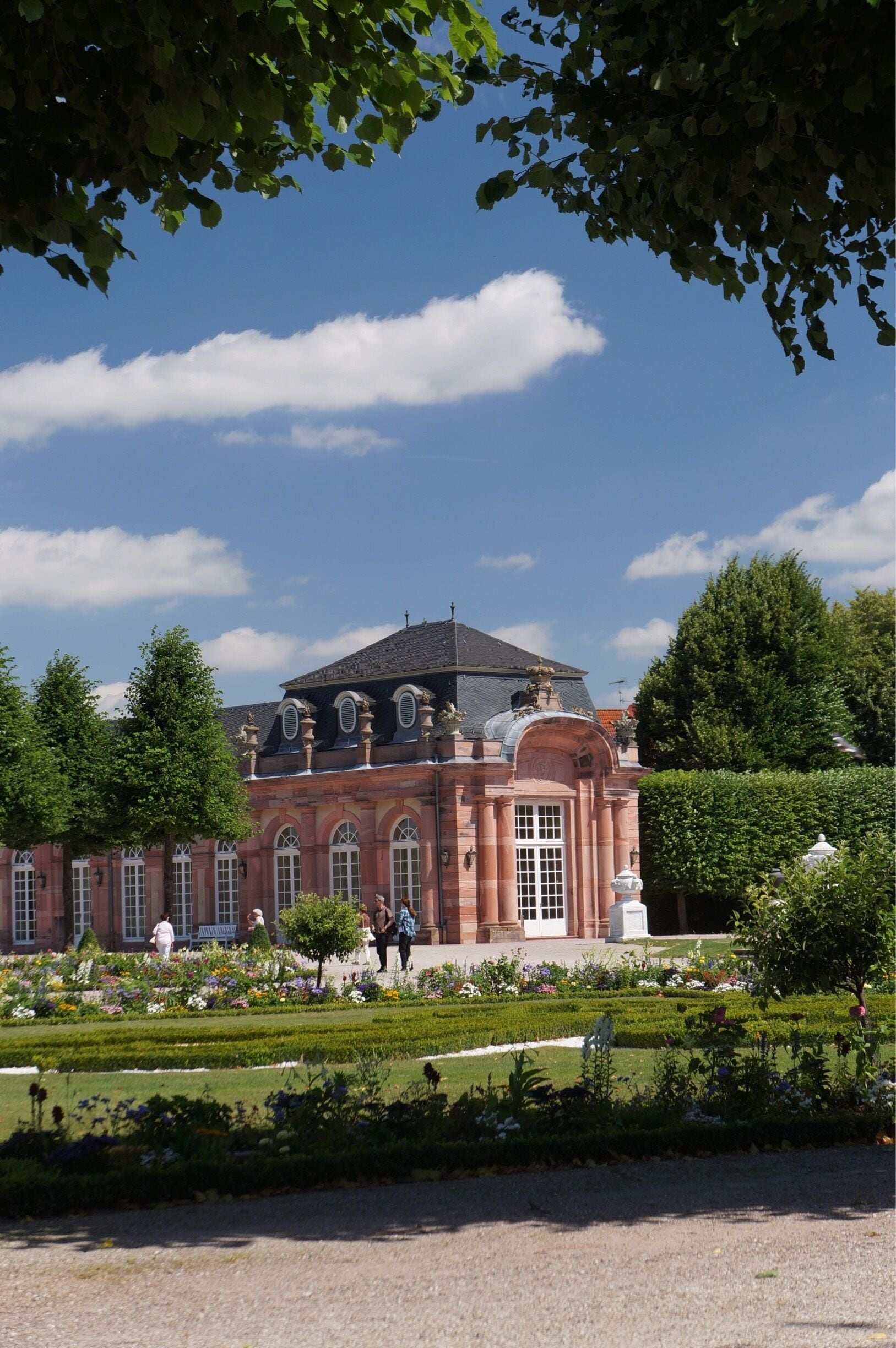 A large, historic building with a red facade and arched windows, surrounded by a well-manicured garden and trees.