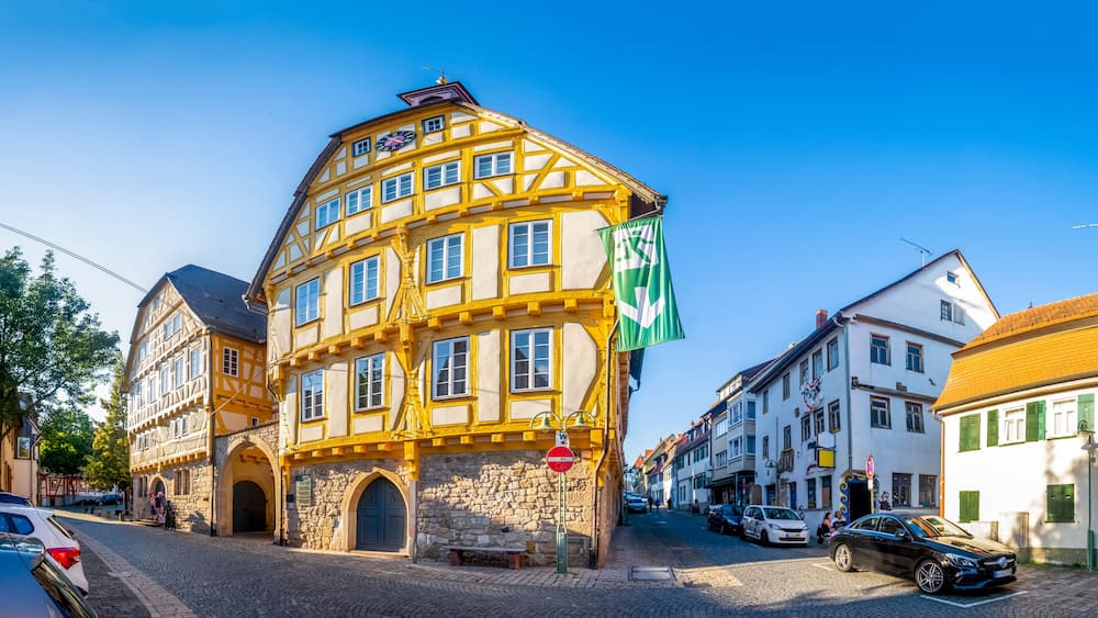 A historic yellow and white building with a clock, cobblestone street, and European flags.
