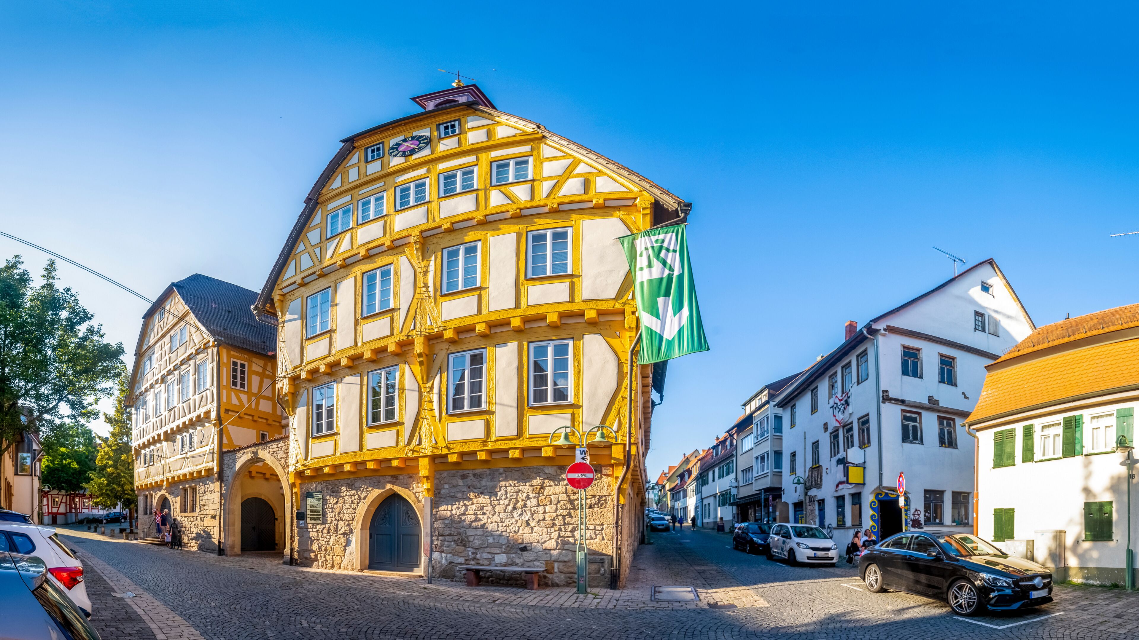 A historic yellow and white building with a clock, cobblestone street, and European flags.
