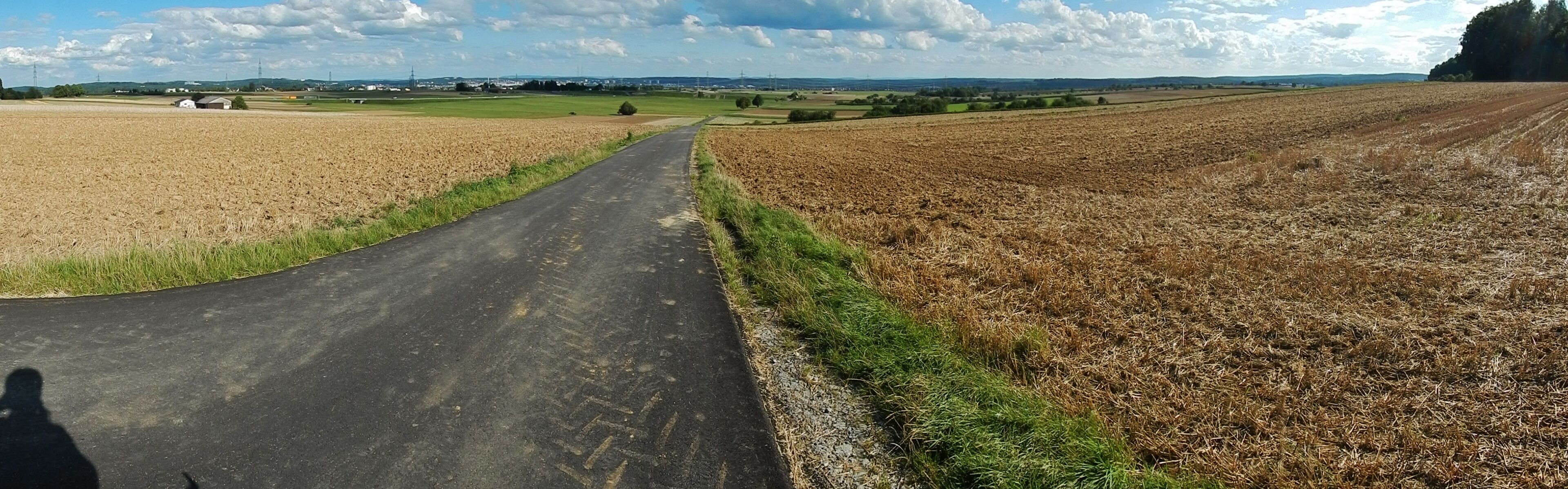 A rural road winding through fields, with a clear sky and distant buildings.