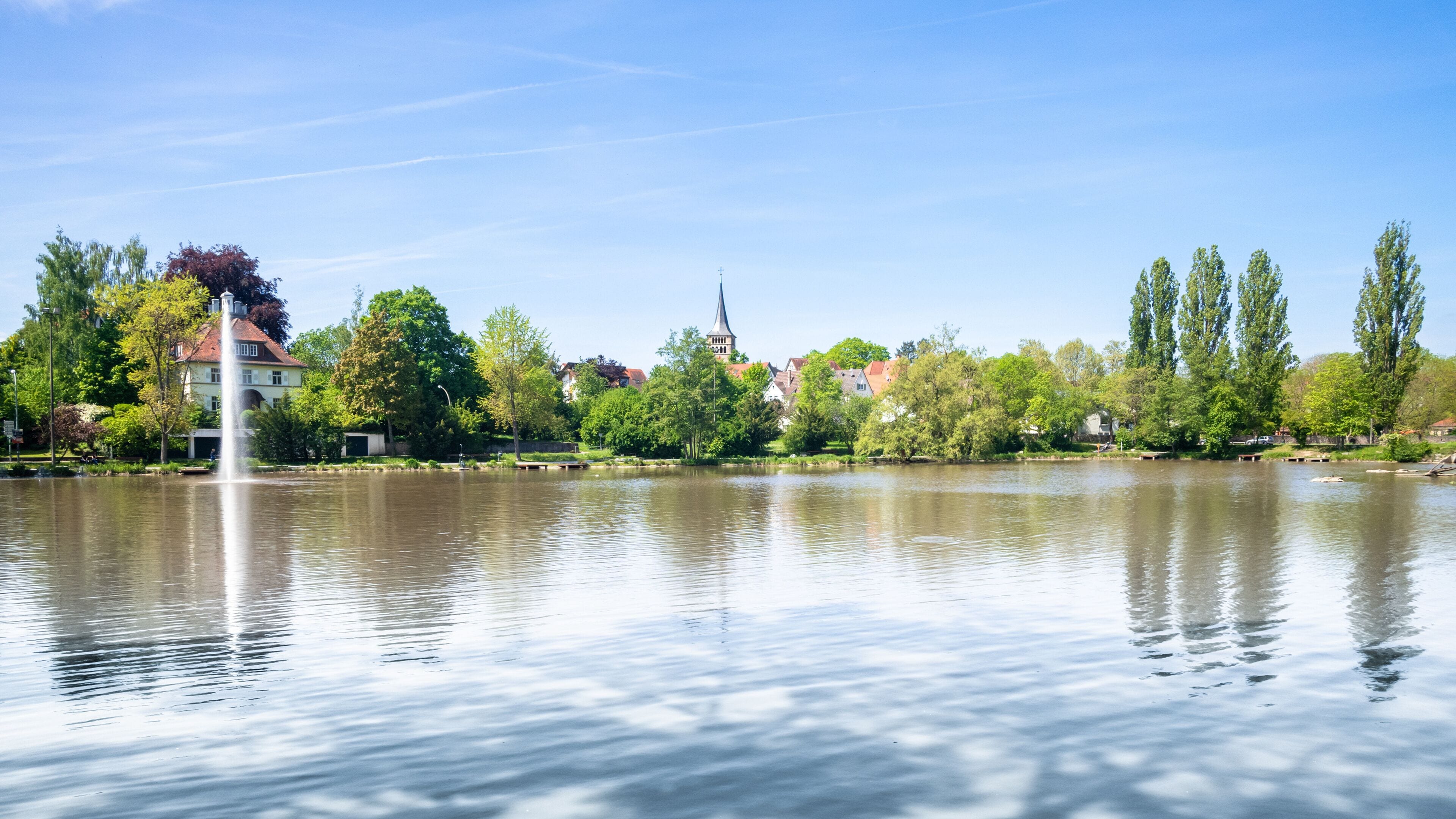 Ein ruhiger See mit einem Brunnen, umgeben von Bäumen und Gebäuden, darunter eine Kirche mit einem Kirchturm.