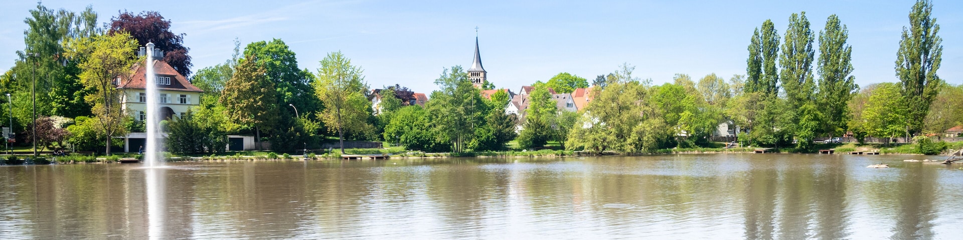 Un lago tranquilo con una fuente, rodeado de árboles y edificios, incluyendo una iglesia con una aguja.