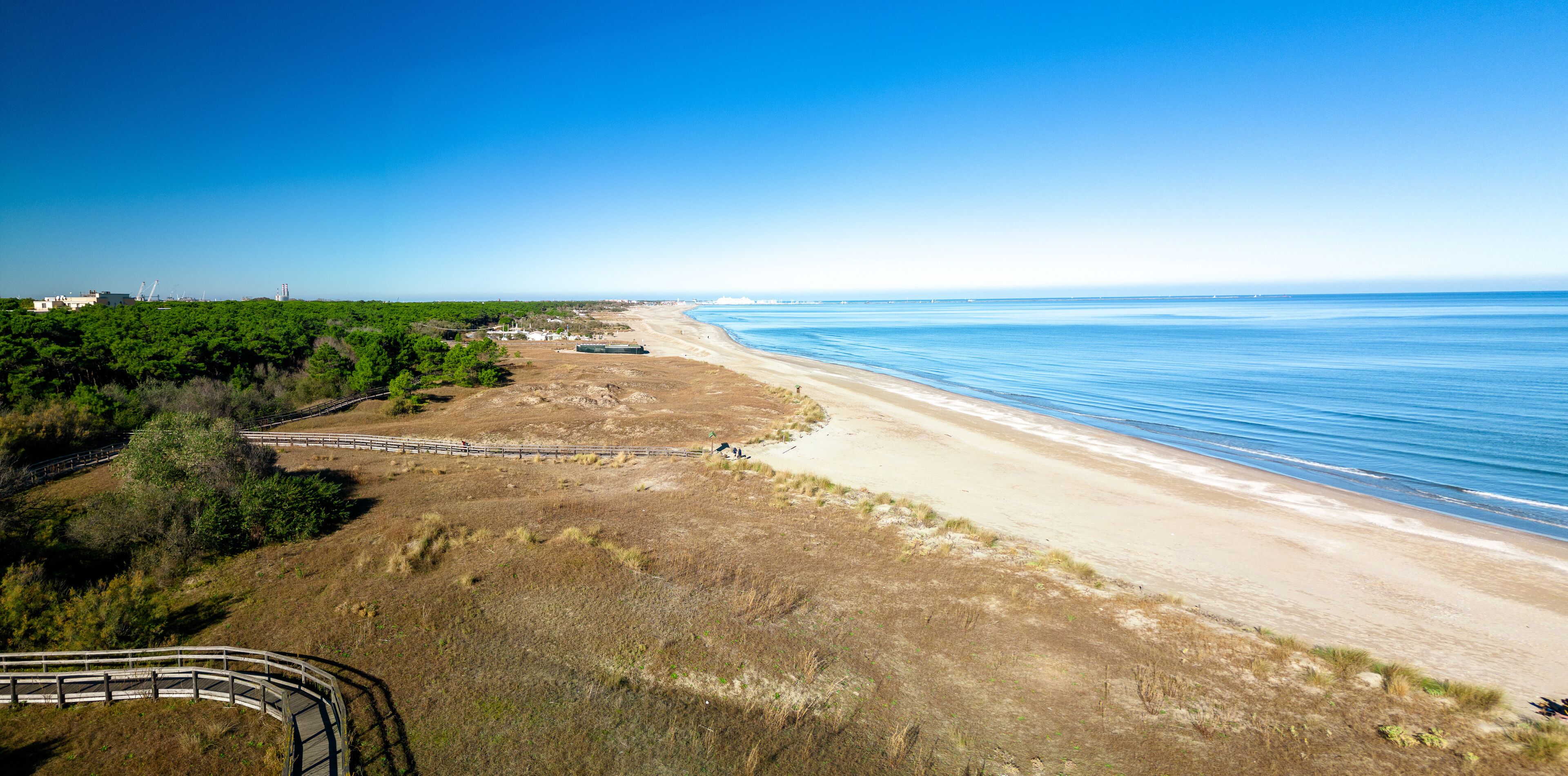 A coastal landscape with a sandy beach, a wooden boardwalk, and a clear blue sky.