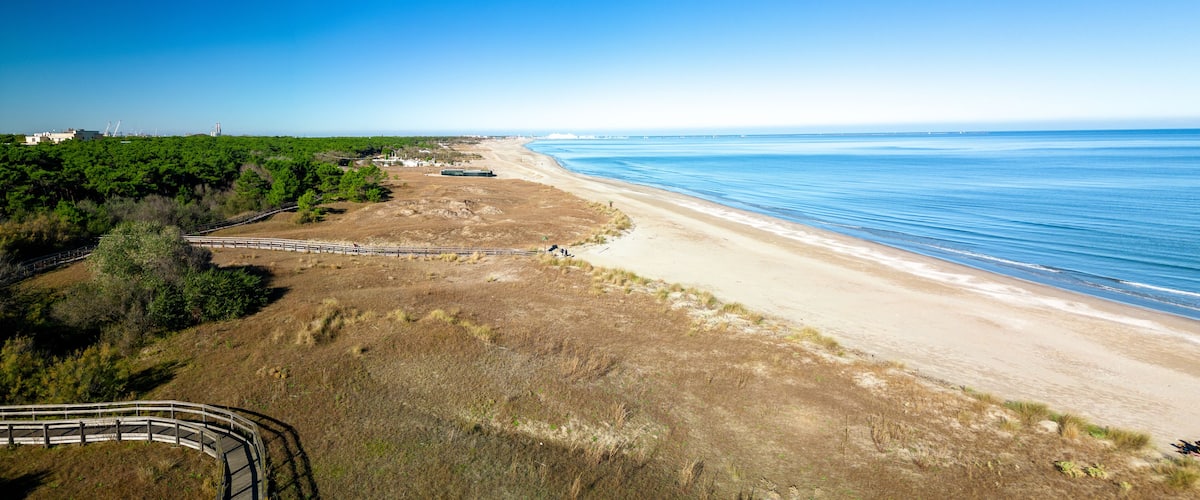 A coastal landscape with a sandy beach, a wooden boardwalk, and a clear blue sky.