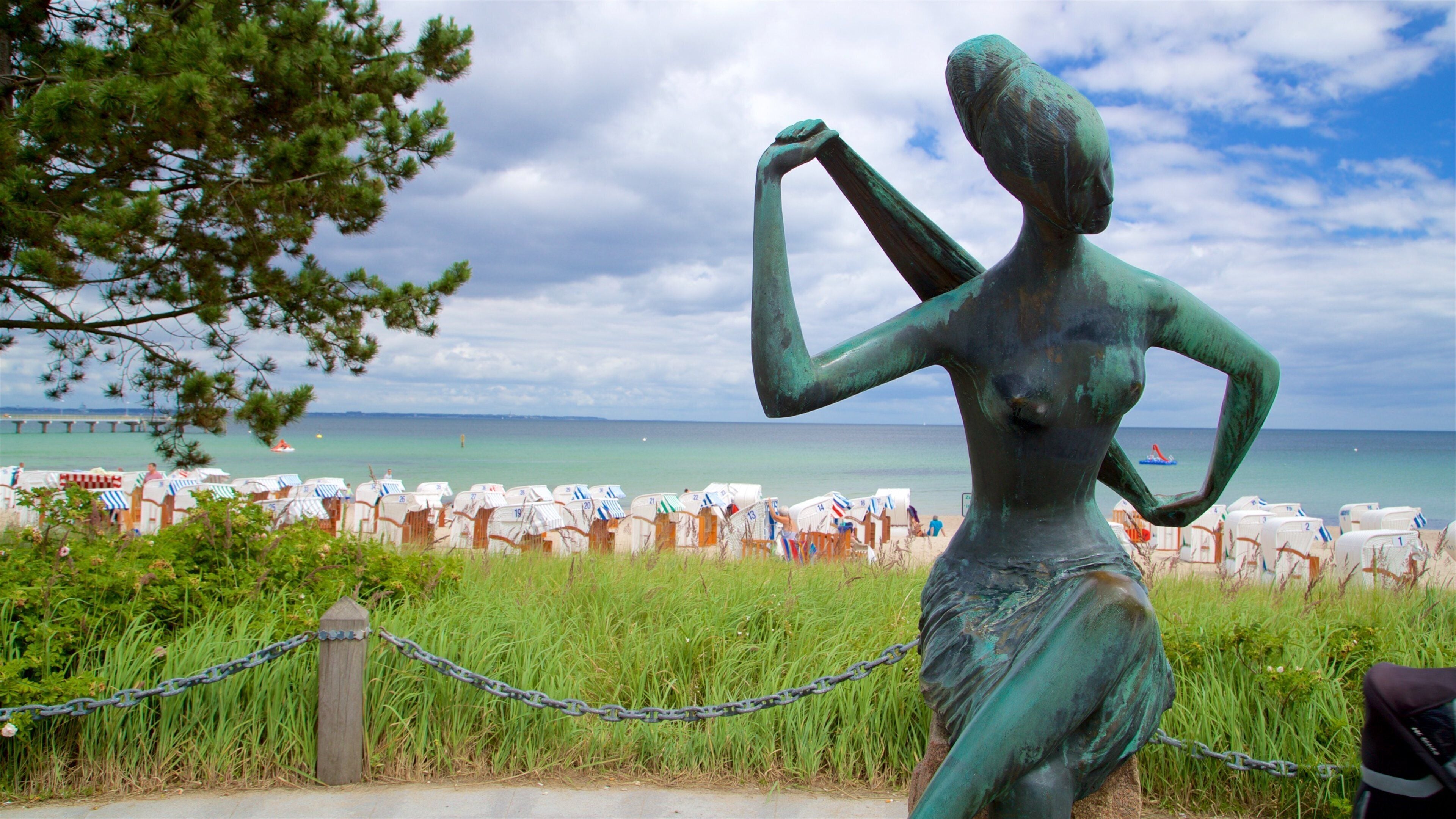 A bronze statue of a woman by the sea, with beach chairs and a pier in the background.