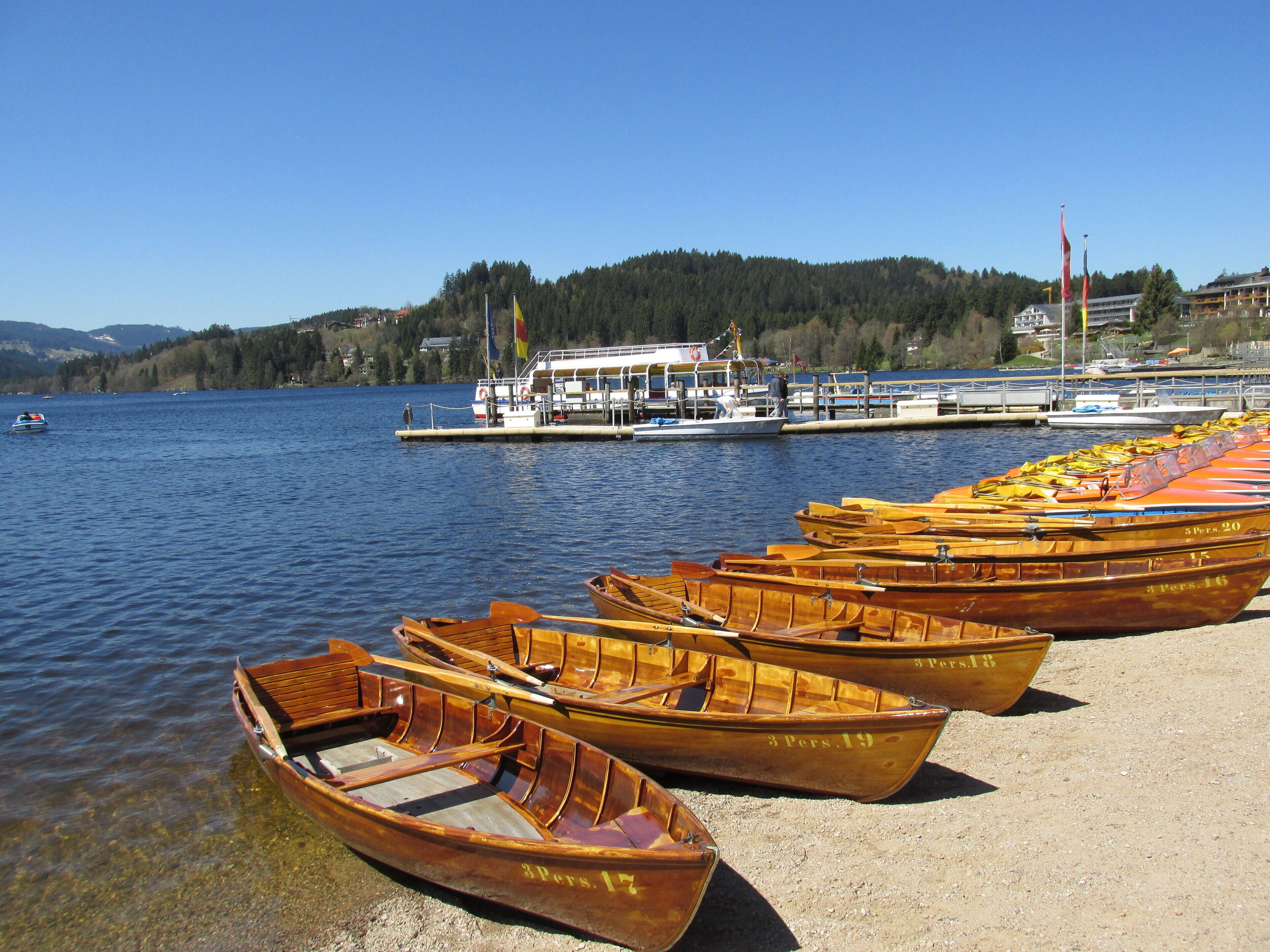 Wooden rowboats lined up on a sandy shore by a calm lake, with a boat dock and buildings in the background.