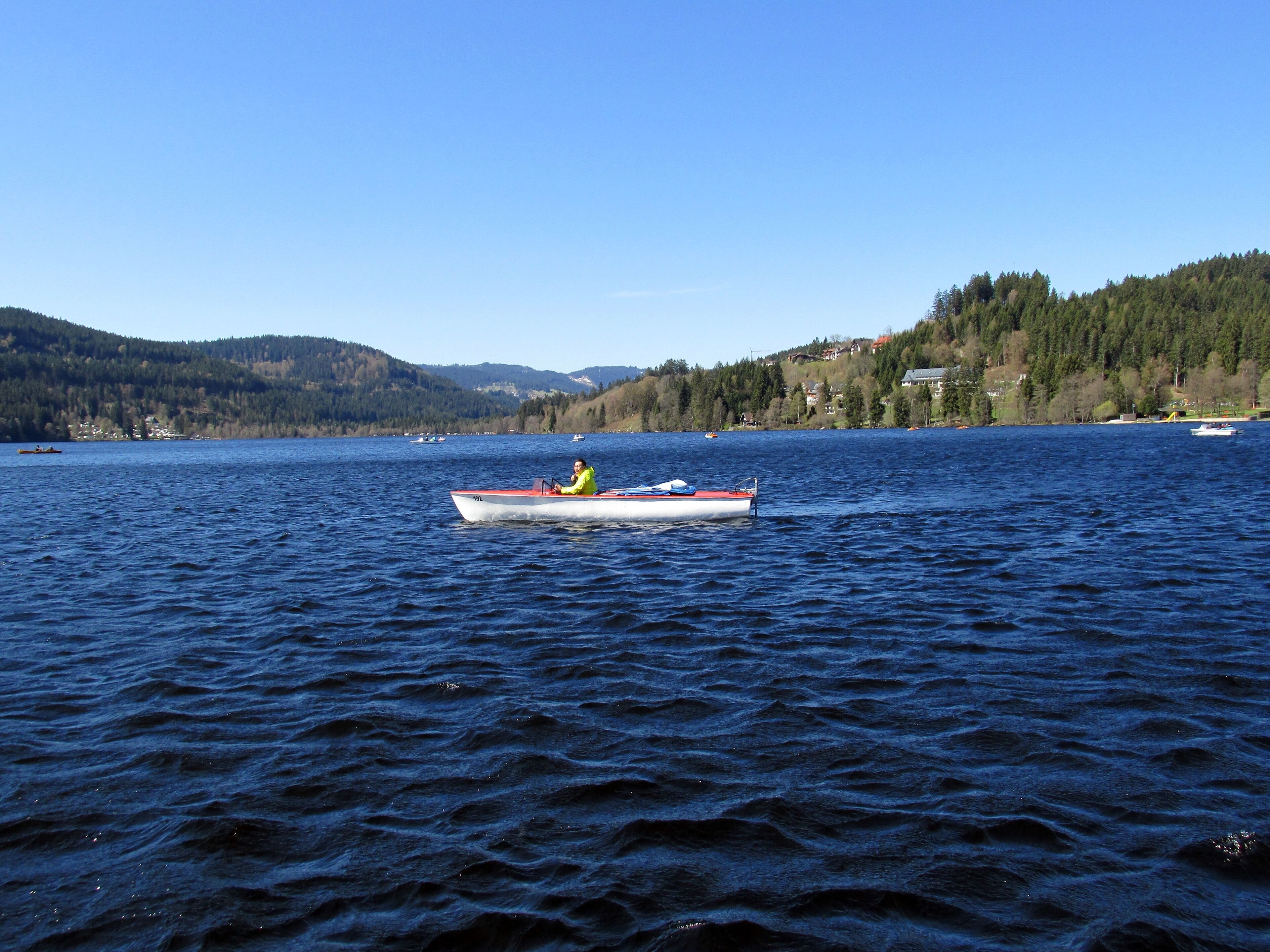 A calm lake with a boat in the center, surrounded by forested hills and houses in the background.