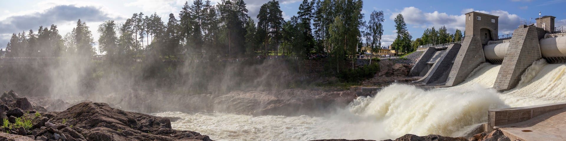 A dam with water cascading over rocks, a bridge, and a forest in the background.