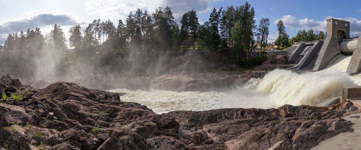 A dam with water cascading over rocks, a bridge, and a forest in the background.