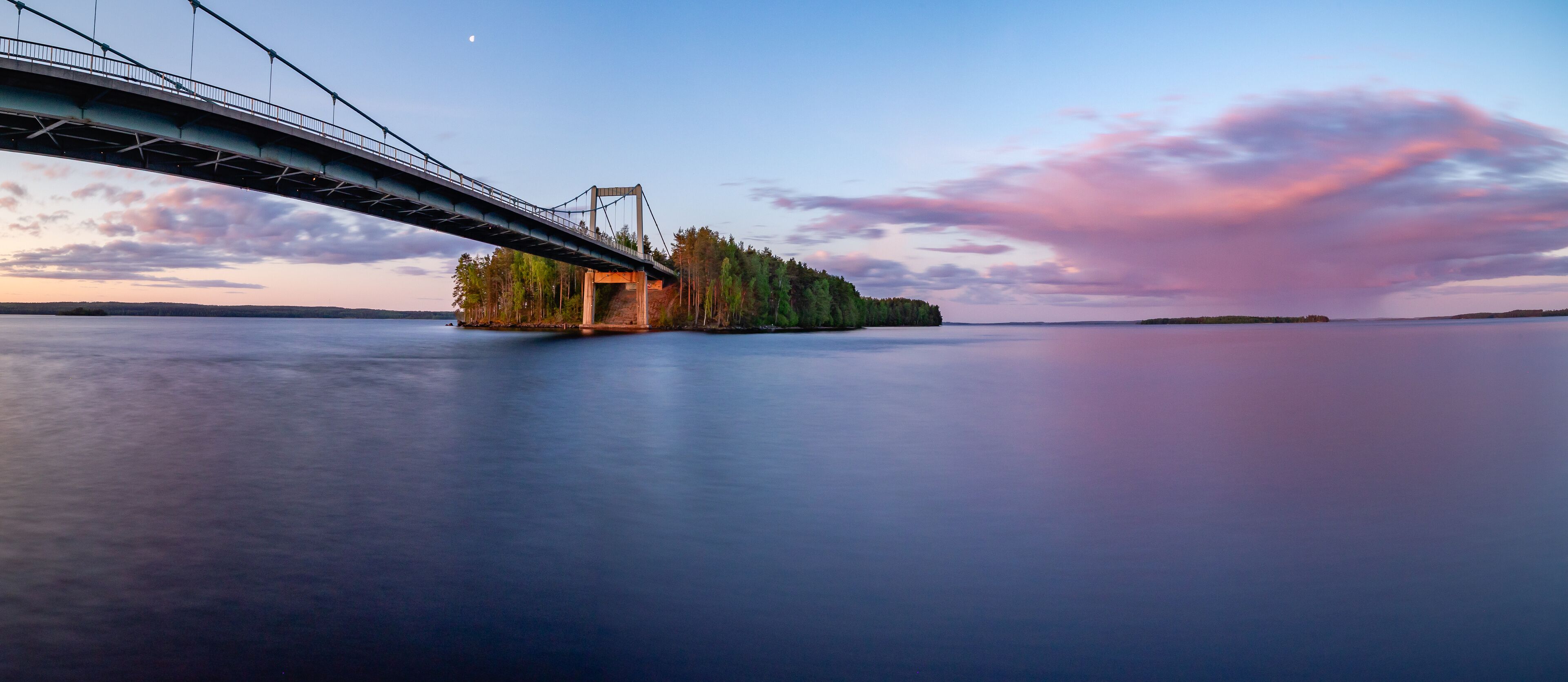 A bridge over calm water, a forested island, and a sunset sky.