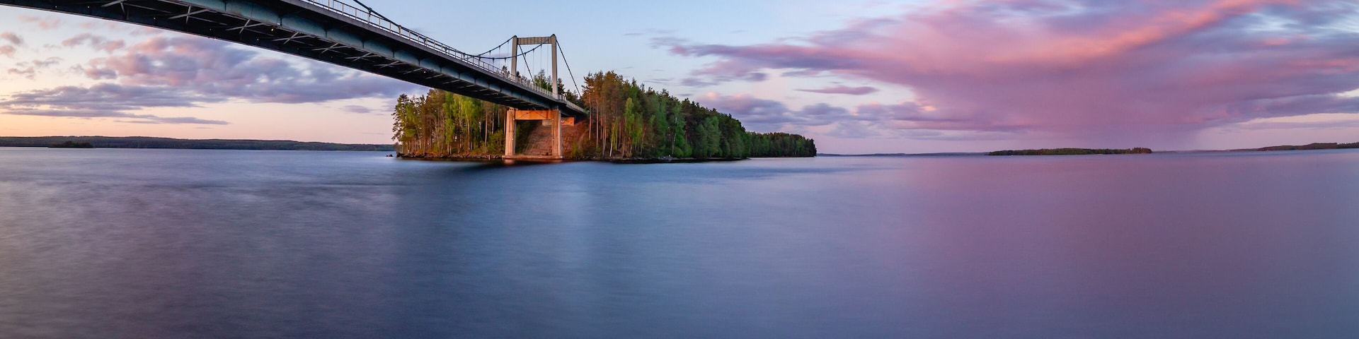 A bridge over calm water, a forested island, and a sunset sky.