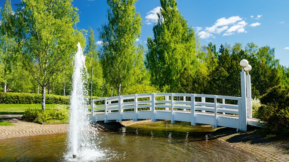 A beautiful park with a pond, wooden bridge and fountain in sunny summer day