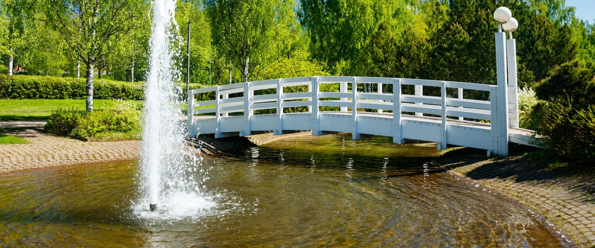 A white bridge over a pond with a fountain in the center.