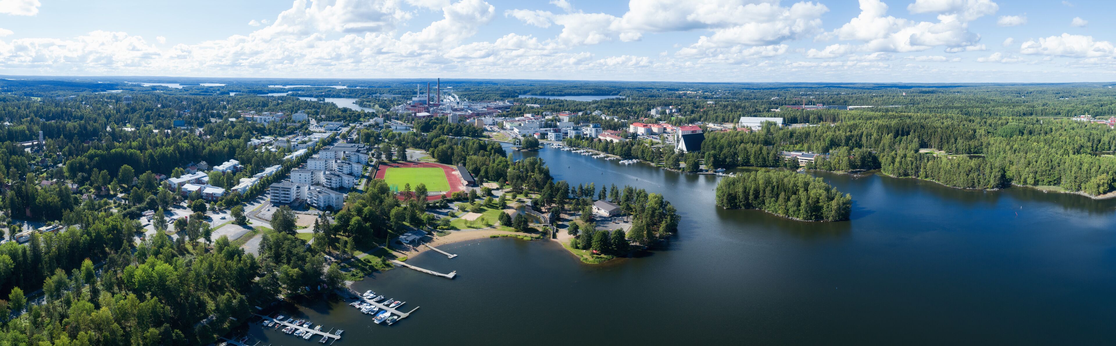 An aerial view of a lakeside town with a marina, a sports field, and surrounding forests.