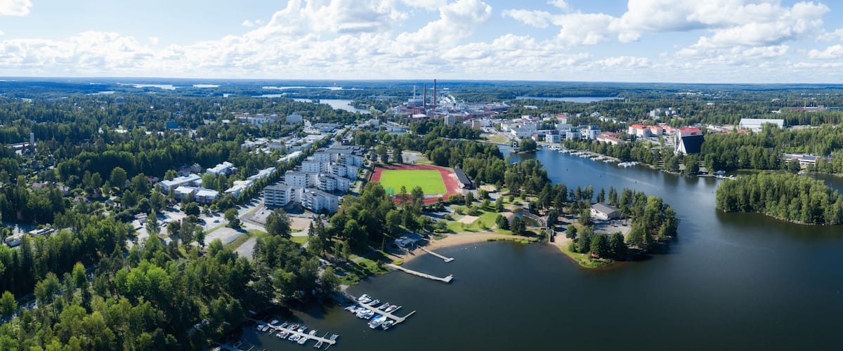 An aerial view of a lakeside town with a marina, a sports field, and surrounding forests.