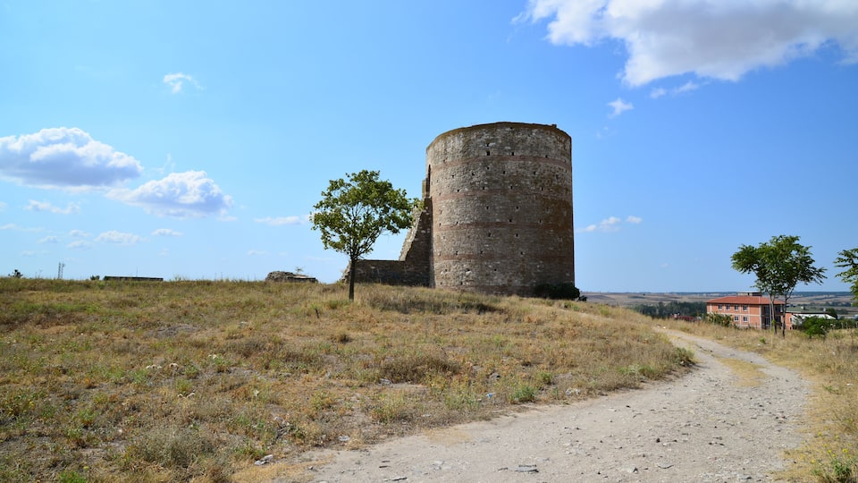 A historic stone tower with a dirt path leading to it, surrounded by open fields and a clear sky.