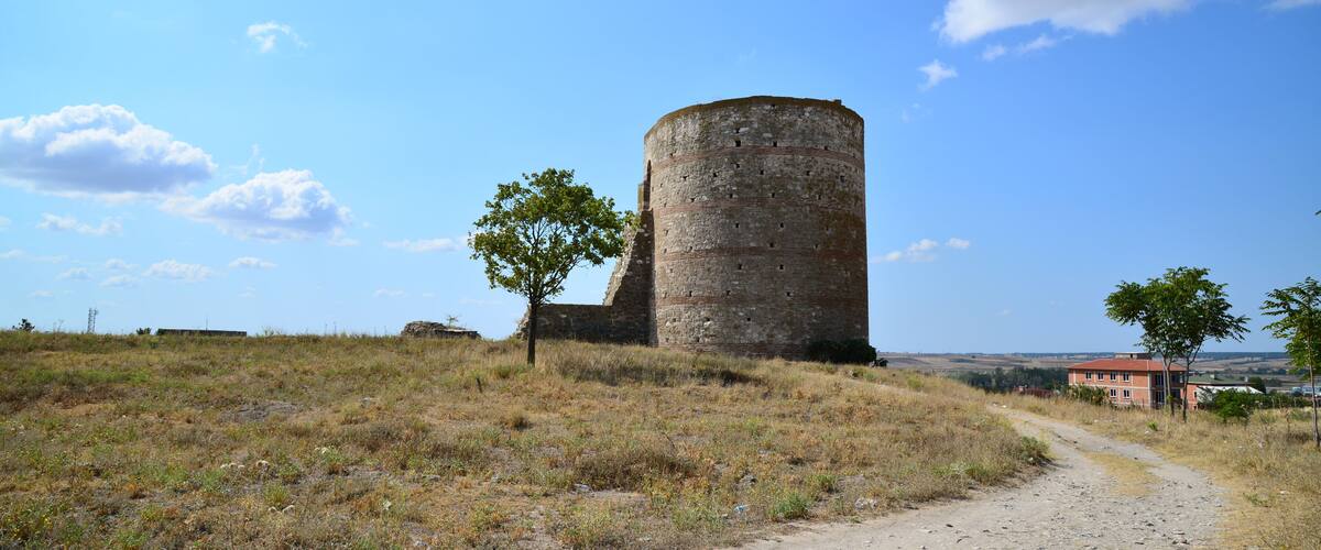 A historic stone tower with a dirt path leading to it, surrounded by open fields and a clear sky.