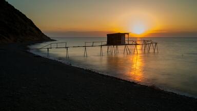 Un atardecer sobre un mar tranquilo con un muelle que se adentra en el agua y una costa rocosa al fondo.