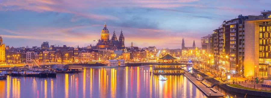 Een stadsgezicht in de schemering met een opvallende kathedraal, een rivier die lichtjes weerspiegelt, en moderne gebouwen.