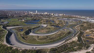 Vista aérea de una pista de carreras serpenteante con múltiples curvas, rodeada de vegetación y un paisaje urbano de fondo.