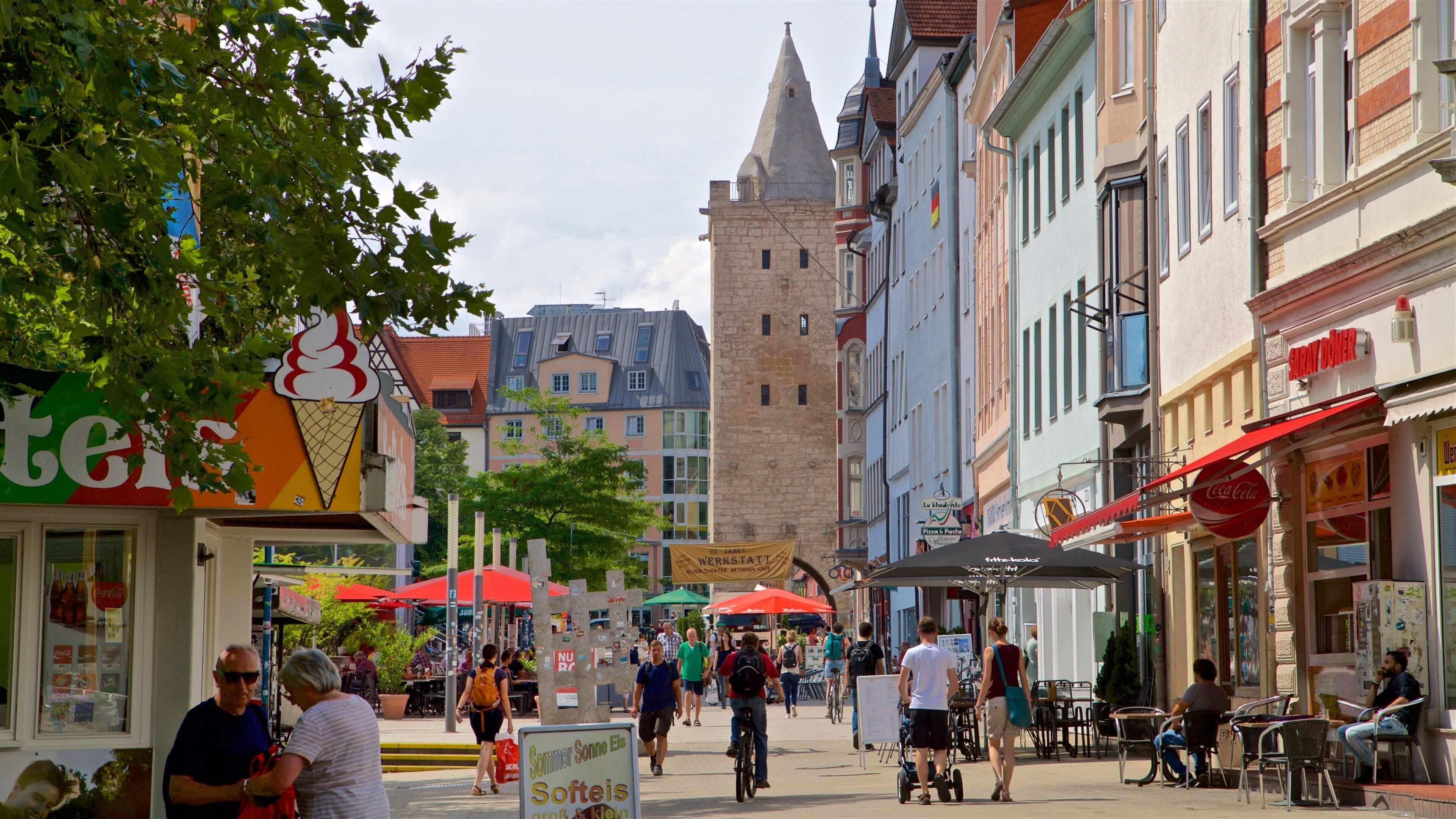 Una animada calle europea con edificios históricos, cafés al aire libre y una torre destacada.