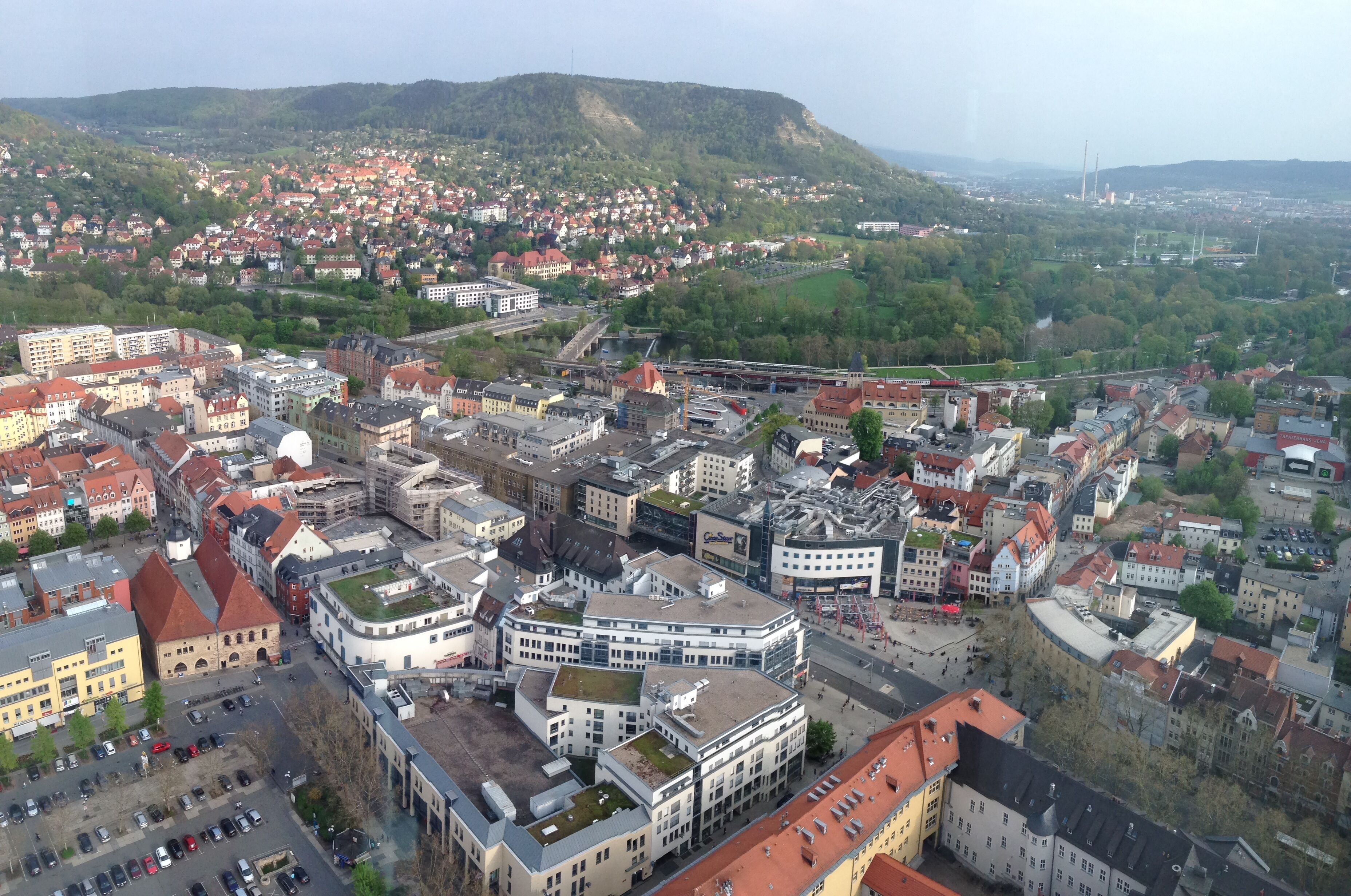 An aerial view of a city with a prominent round building, a railway line, and a hilly background.