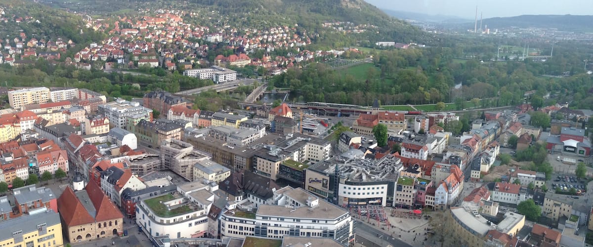 An aerial view of a city with a prominent round building, a railway line, and a hilly background.