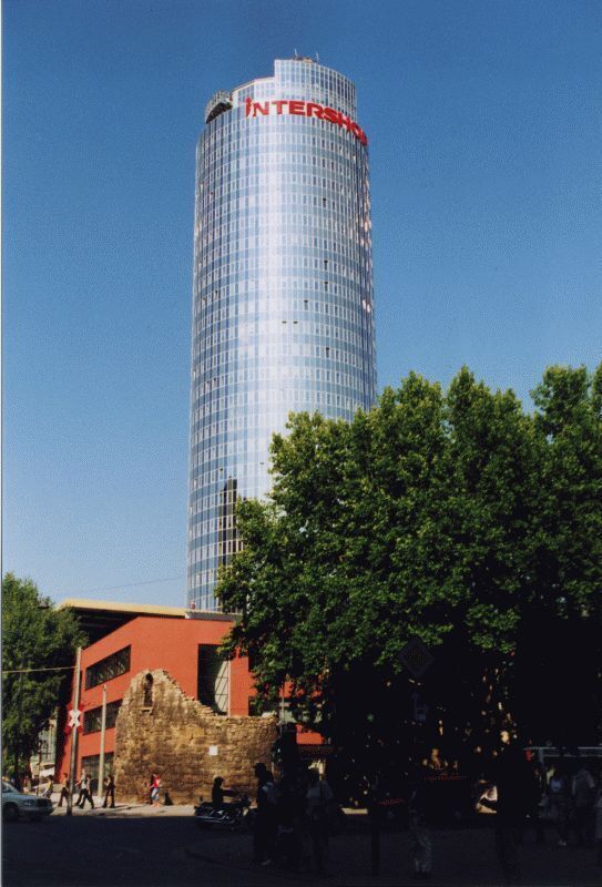 A tall glass building with the word "INTERSHOP" on top, surrounded by trees and a historic stone structure.