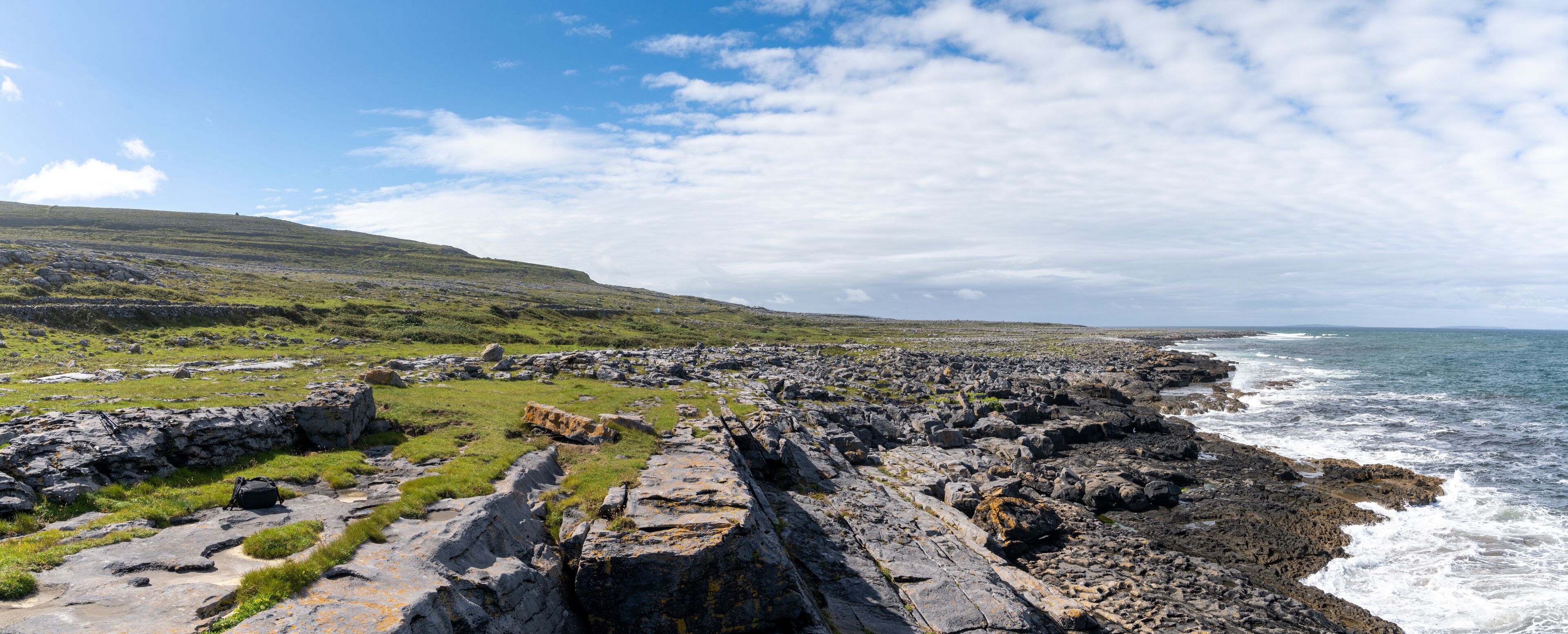 A rocky coastline with waves crashing against the shore, a grassy hill in the background, and a clear sky.