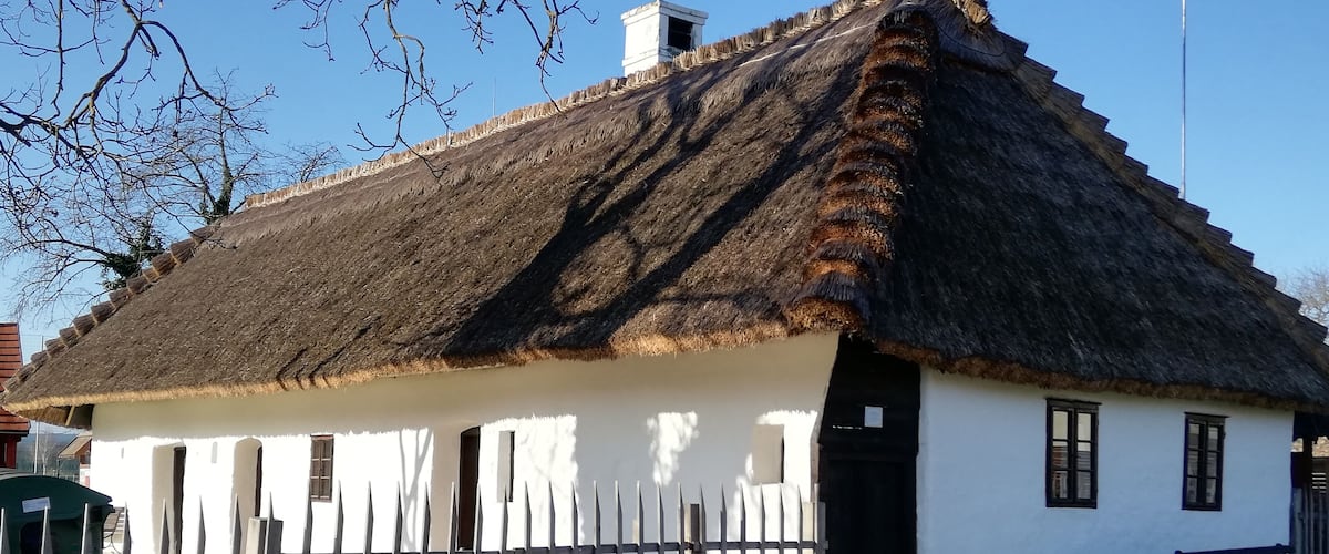 A traditional thatched-roof house with a white exterior, a wooden fence, and a bench nearby.